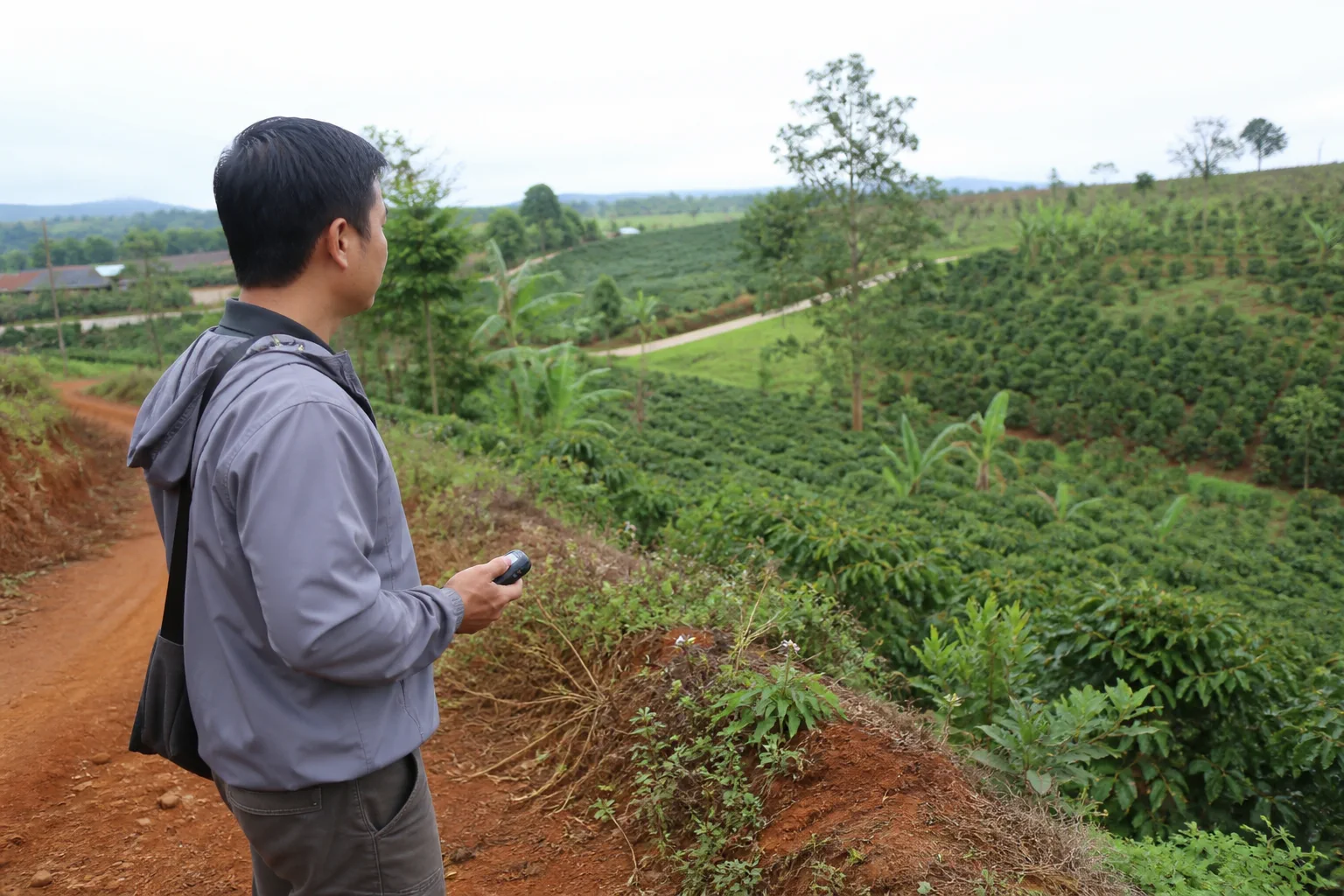 Người khảo sát thực địa tại Đắk Nông đứng bên bờ khu đất nông nghiệp, dùng thiết bị ghi nhận vị trí và quan sát các mốc cảnh quan tự nhiên xung quanh để chuẩn bị biên bản khảo sát