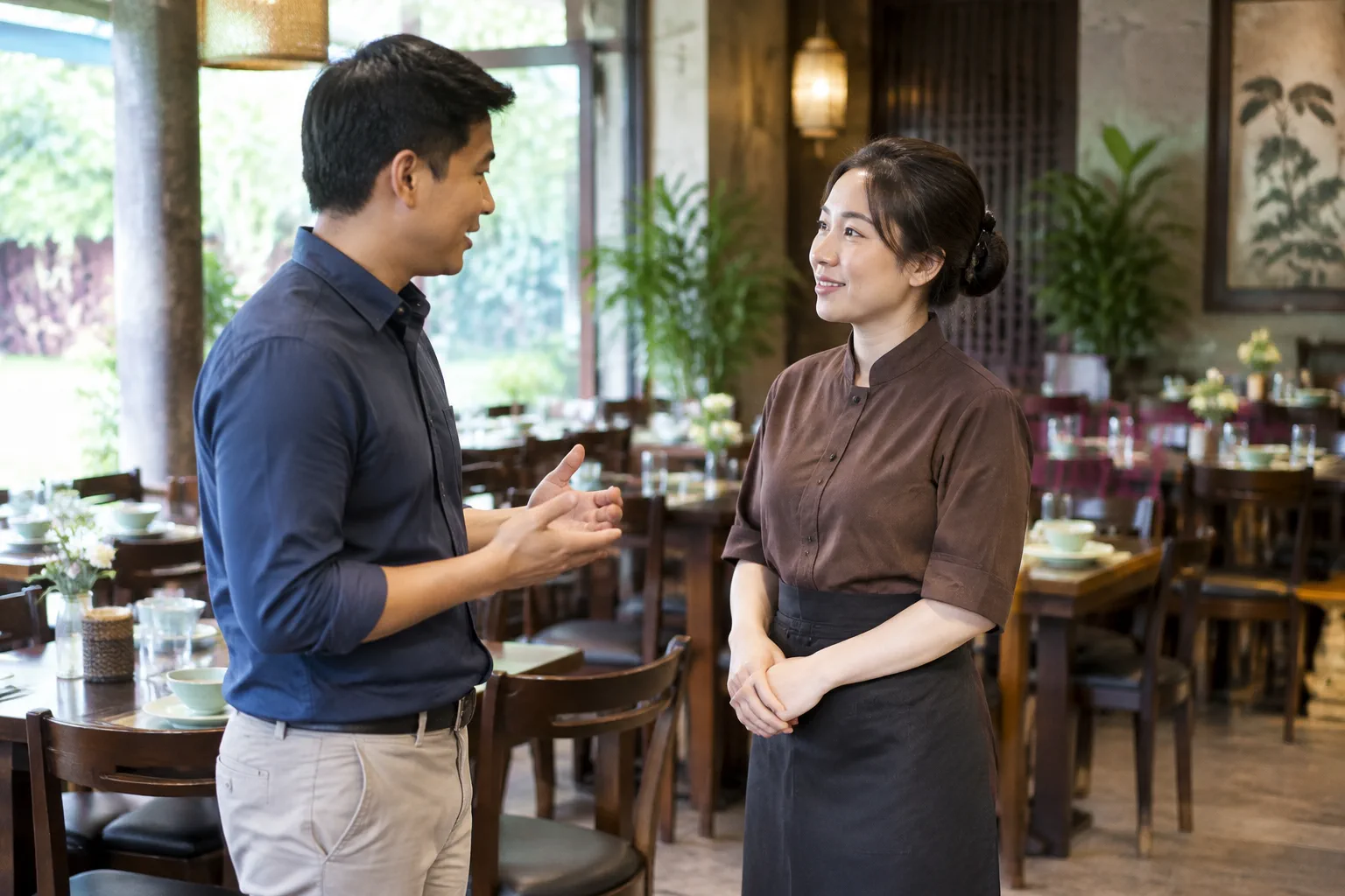 Trưởng đoàn và nhân viên nhà hàng trao đổi trực tiếp về thời gian dùng bữa và nhu cầu không gian cho nhóm khách trong khu vực tiếp khách, thể hiện bước chuẩn bị trước chuyến đi.