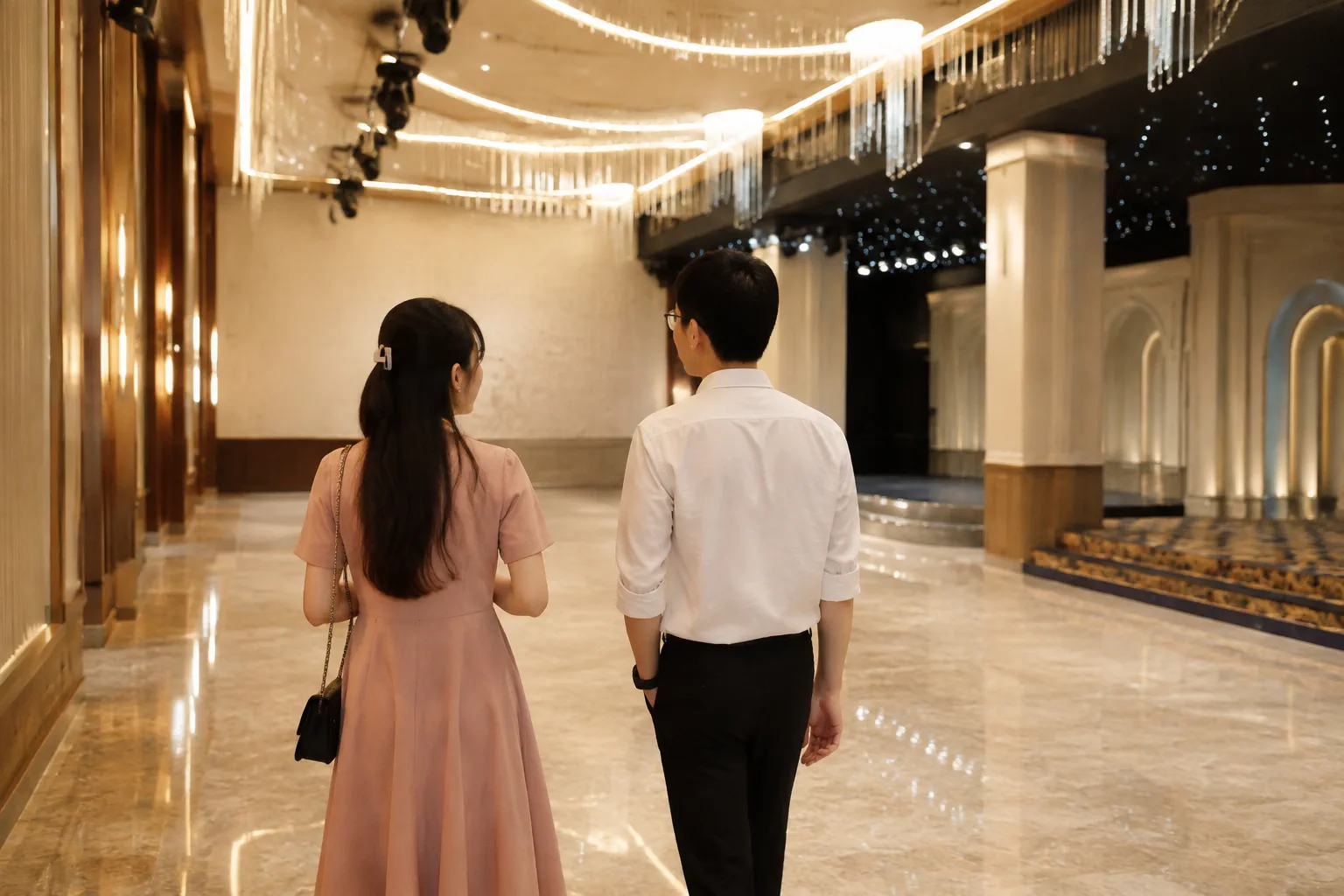 Góc nhìn từ phía sau một cặp đôi Việt Nam đang quan sát lối di chuyển và khu vực bố trí sự kiện trong nhà hàng tiệc cưới, thể hiện việc đánh giá không gian thực tế trước khi lựa ch
