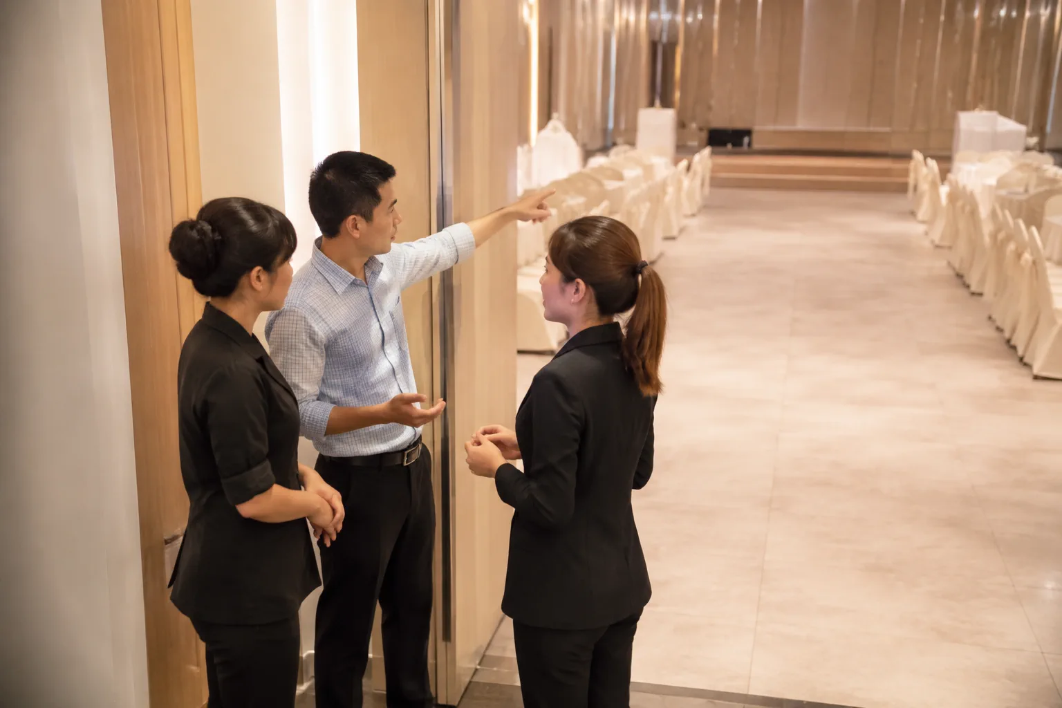Người đại diện gia đình trao đổi riêng với nhân viên nhà hàng và người phụ trách chương trình bên cạnh lối đi sảnh tiệc, thể hiện phối hợp các thay đổi trong ngày cưới